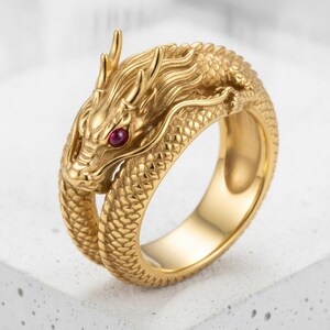 May include: A gold-toned ring shaped like a coiled dragon with a textured scale pattern covering the band. The dragon head features a small, round, deep red gemstone set as an eye, with detailed facial features including horns and whiskers.