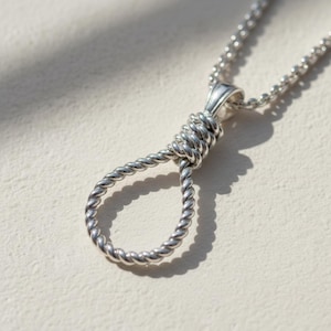 May include: Silver pendant necklace with a noose design. The pendant has a twisted rope texture and hangs from a fine chain. The necklace is shown against a neutral backdrop, emphasizing the detailed craftsmanship of the piece.