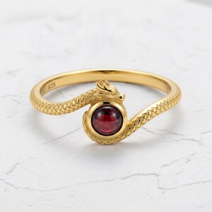 May include: A gold-toned ring featuring a textured serpent design that wraps around a central, round, deep red gemstone. The band is marked with the number 925 on the inner surface, indicating sterling silver construction with a gold-coloured finish.