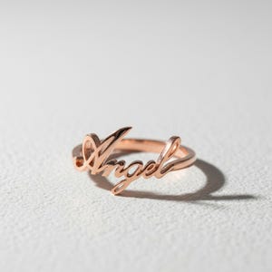 以下が含まれることがあります： 筆記体でAngelという文字がデザインされた、ローズゴールドカラーの金属製リング。リングは光沢のある仕上げで、文字部分がジュエリーの焦点となるような連続したバンドデザインになっています。