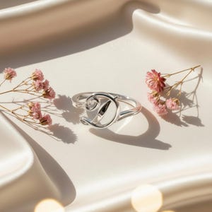 Può includere: Un anello d'argento con la lettera "D" in corsivo. L'anello è posto su uno sfondo di raso bianco, con fiori rosa su entrambi i lati. Un gioiello delicato ed elegante.