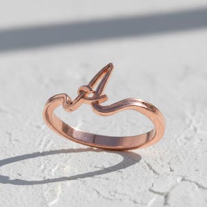 Może przedstawiać: Pierścionek z różowego złota z literą "A" w ozdobnej czcionce. Pierścionek ma delikatny, minimalistyczny design z gładkim, polerowanym wykończeniem. Litera "A" jest centralnym punktem, dodając biżuterii osobistego charakteru.