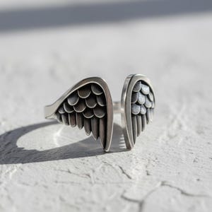 May include: A silver-colored ring with an open design, featuring two stylized wing shapes. Each wing has detailed feather patterns. The ring is set against a white, textured surface, with a shadow cast to the left.