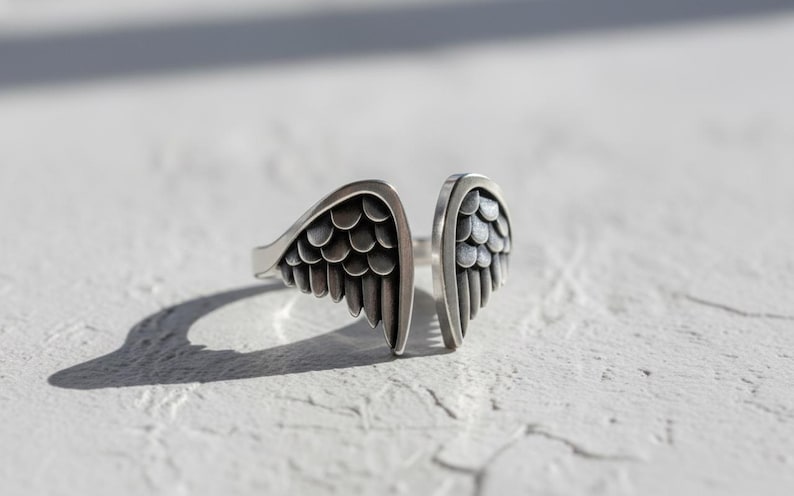 May include: A silver-colored ring with an open design, featuring two stylized wing shapes. Each wing has detailed feather patterns. The ring is set against a white, textured surface, with a shadow cast to the left.