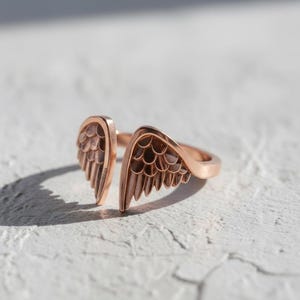 May include: A rose gold ring with an open design featuring two detailed wing shapes. The ring is set against a textured white surface, with a shadow cast to the left. The jewelry piece is a symbol of freedom and elegance.