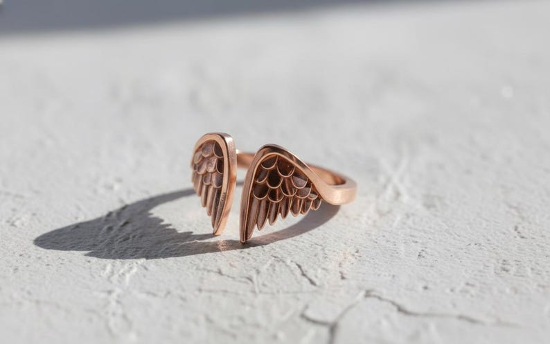 May include: A rose gold ring with an open design featuring two detailed wing shapes. The ring is set against a textured white surface, with a shadow cast to the left. The jewelry piece is a symbol of freedom and elegance.