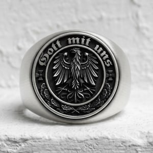 May include: A silver-toned metal ring featuring a circular face with an embossed eagle emblem at the center. The text "Gott mit uns" is inscribed along the upper edge of the circular border, flanked by small cross symbols and oak leaf motifs.
