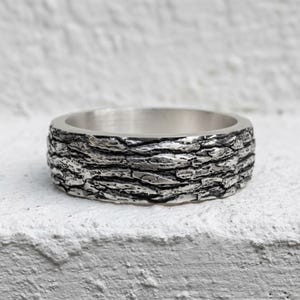 May include: A silver ring featuring a highly detailed, textured exterior surface that mimics the appearance of natural tree bark. The band has a smooth, polished interior finish and a dark, oxidized patina within the deep crevices of the pattern.