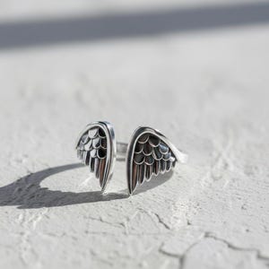 May include: A silver-toned ring featuring an open design with two detailed wing shapes. The wings have a textured, feathered appearance, and the ring is set against a white, textured surface. The ring's design suggests a theme of freedom or spirituality.