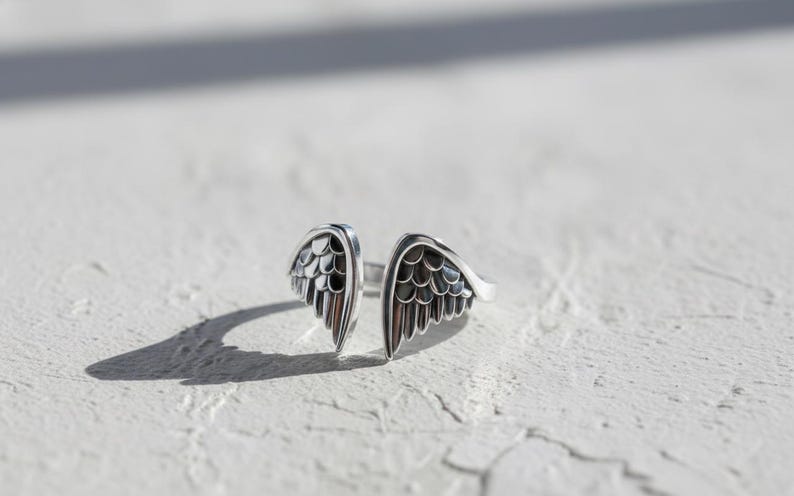 May include: A silver-toned ring featuring an open design with two detailed wing shapes. The wings have a textured, feathered appearance, and the ring is set against a white, textured surface. The ring's design suggests a theme of freedom or spirituality.
