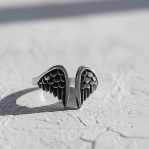 May include: A silver-toned ring with an open design, featuring two detailed wing shapes. The wings have a textured, feathered appearance, and the ring is set against a white, textured surface. The ring's design suggests a theme of freedom or spirituality.
