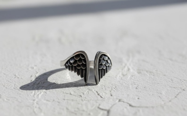 May include: A silver-toned ring with an open design, featuring two detailed wing shapes. The wings have a textured, feathered appearance, and the ring is set against a white, textured surface. The ring's design suggests a theme of freedom or spirituality.