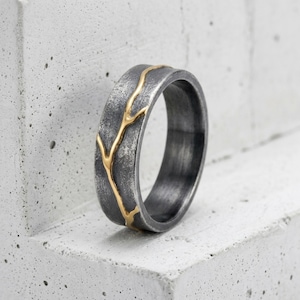May include: A band ring featuring a dark, oxidized grey finish with a textured, rugged surface. A single, thin, polished gold-colored line runs vertically across the band, resembling a branching vein or crack, creating a contrast in color and texture.