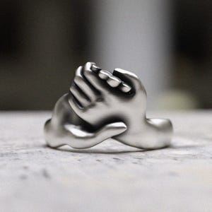Puede incluir: Un anillo de plata con dos manos entrelazadas, simbolizando la unidad y el amor.