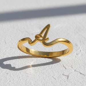 Può includere: Un anello color oro con la lettera "A" in un design corsivo. L'anello ha una finitura liscia e lucida. L'anello è posto su una superficie bianca. Il design dell'anello è una linea continua.
