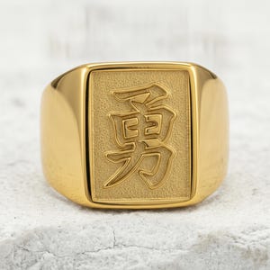Könnte beinhalten: Ein goldfarbener Siegelring mit rechteckiger Front und texturiertem Hintergrund. In der Mitte des Rings befindet sich ein erhabenes, poliertes japanisches Kanji-Zeichen für Mut, das auf einer matten, vertieften rechteckigen Fläche liegt.