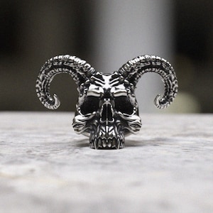 Può includere: Un anello in argento a forma di teschio con corna. Il teschio è dettagliato con denti affilati e corna.