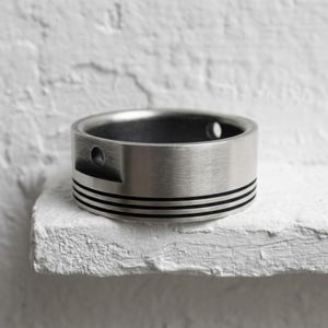 Könnte beinhalten: Ein breiter Ring aus gebürstetem, silberfarbenem Metall mit einer flachen Außenfläche, die von drei horizontalen, schwarzen, eingravierten Linien umrundet wird. Der Ring weist an der Seite eine kleine, kreisförmige Vertiefung auf.