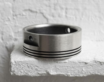 May include: A wide, brushed silver-tone metal ring featuring a flat outer surface with three horizontal black engraved lines circling the band. The ring includes a small, circular indentation on the side, creating a distinct, industrial-inspired design.
