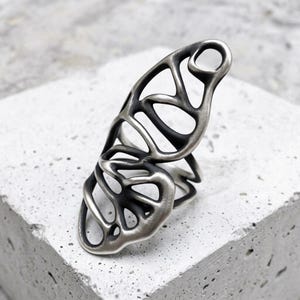 May include: A silver-toned ring featuring an elongated, openwork design with organic, flowing lines and dark recessed areas. The metal surface has a brushed finish, creating a contrast between the polished raised edges and the darkened interior spaces.