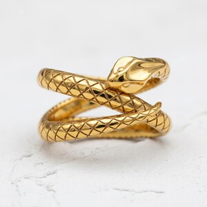 May include: A gold-colored snake ring with a textured, scale-like pattern. The snake's head is raised, and the body wraps around the finger. The ring is a piece of jewelry.