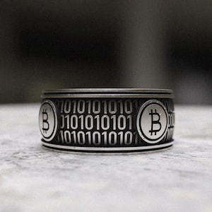 May include: A silver ring with a black band featuring the Bitcoin symbol and binary code.