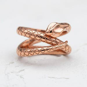 May include: A rose gold snake ring with a textured, scale-like pattern. The snake's head is raised, and the body wraps around the finger in a spiral design. The ring is set against a white background.