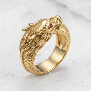May include: A gold-toned ring featuring a detailed dragon head design with flowing mane textures and scale patterns along the band. The dragon eye is accented with a small, round, deep red gemstone set into the metal surface of the head.