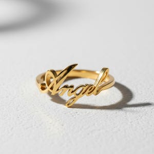 Può includere: Un anello in metallo color oro con la parola Angel scritta in un carattere corsivo. L'anello presenta una finitura lucida e una fascia continua che si collega alla scritta, creando un gioiello con una superficie liscia e riflettente.