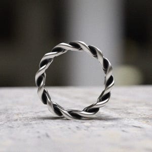 May include: A silver-colored ring with a twisted rope design. The ring is circular and appears to be made of metal. The twisted pattern creates a textured surface. The ring is displayed on a light-colored surface.