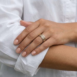 May include: A silver and gold ring with an intricate, repeating swirl design. The ring is worn on a person's finger, with a white shirt in the background. The ring's design is detailed and eye-catching, suitable for everyday wear or special occasions.