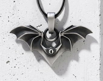 May include: A silver-toned metal pendant shaped like bat wings with a central eye motif. The pendant features a brushed finish and is suspended from a matching silver-toned snake chain. The design includes a circular bail connecting the wings.