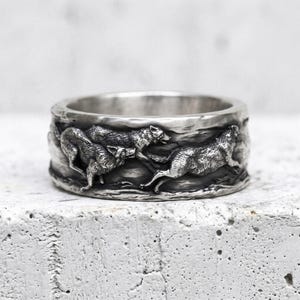May include: A silver-toned metal ring featuring a detailed, raised relief design of multiple wolves in motion. The band has an oxidised finish that highlights the texture of the fur and the surrounding landscape, creating a high-contrast appearance.