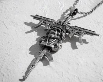 May include: A silver-toned metal pendant necklace featuring a central skull wearing a crown, positioned at the intersection of two horizontal submachine guns and one vertical assault rifle, all suspended from a matching silver-toned chain link.