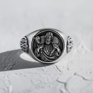 May include: Silver-toned ring featuring a detailed relief of a figure carrying a lamb. The ring has an oval face with a circular central design and decorative side accents. The ring is set against a textured white surface.