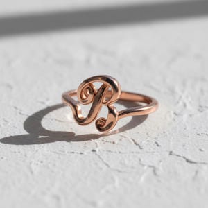 May include: A rose gold ring with the cursive letter "B" as its main design. The ring is set against a textured white surface, with a shadow to the left. The ring's design is delicate and elegant, suitable for everyday wear.