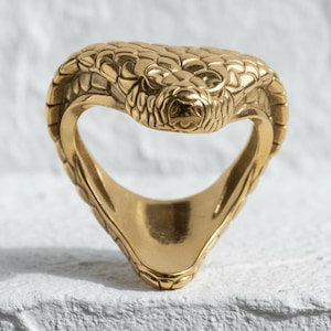 Op de afbeelding: Een goudkleurige ring in de vorm van een cobra met een gestructureerd, schubachtig patroon op de kop en de kap. De ringband vormt het onderlichaam van de slang, wat zorgt voor een doorlopend ontwerp in een open lus met een gepolijste afwerking.