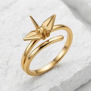 May include: A gold-toned ring featuring a stylized origami crane design. The band is open-ended, with the crane motif positioned at the top. The metal has a smooth, polished finish, reflecting light across the wings and body of the bird shape.