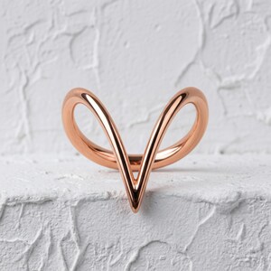 Peut inclure: Une bague en or rose composée d'un anneau lisse et continu qui se courbe vers le haut pour former une pointe en V au centre. Le métal présente une finition polie et réfléchissante, créant deux boucles symétriques de chaque côté.