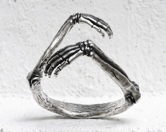 May include: A silver-toned metal bracelet featuring a detailed design of two skeletal hands reaching toward each other. The band is textured to resemble bone, with dark oxidized accents highlighting the anatomical structure of the fingers and wrists.