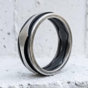May include: A silver-toned metal ring featuring a dual-layered design with a brushed, weathered finish. The outer band has a smooth, polished edge, while the inner section displays a dark, recessed, and angular geometric pattern throughout.