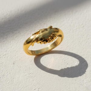 May include: A gold-colored ring with a detailed design of two hands clasped together. The ring is set against a white surface, with a shadow cast by the ring. The ring's design is intricate and the metal has a polished finish.