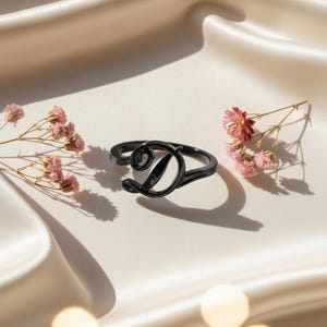 May include: A black ring in the shape of the letter "D" rests on a white satin surface, with pink flowers on either side. The ring is a bold, modern design, perfect for a personalized touch.