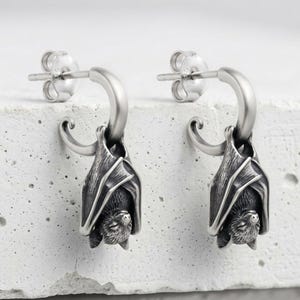 May include: A pair of silver hoop earrings featuring a detailed, three-dimensional bat charm hanging from each hoop. The bats are depicted in a resting, upside-down position with wings wrapped around their bodies and textured fur details.
