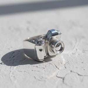 May include: A silver camera-shaped ring, featuring a detailed lens and body. The ring is highly polished, reflecting light and creating highlights and shadows. A unique piece of jewellery.