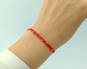 May include: A red string bracelet with gold beads. The bracelet is worn on a person's wrist.
