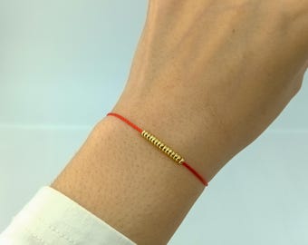 May include: A red string bracelet with a gold bead charm. The bracelet is worn on a person's wrist.
