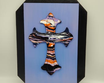 May include: A decorative cross with a marbled design in orange, black, white, and purple hues. The cross is mounted on a light blue background, framed by a black, octagonal border. The artist's signature, "Dawn Ojan," is visible at the bottom.
