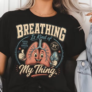 May include: A black t-shirt featuring a graphic of a smiling pair of lungs with a thumbs up and a respiratory mask. The text reads "BREATHING Is Kind of My Thing." and includes "ASSESS. TREAT. EDUCATE." and "RESPIRATORY THERAPIST RT" in a circular design.