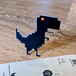 Puede incluir: Una figura de dinosaurio pixelado negro con un ojo rojo, que recuerda a un personaje clásico de videojuego. El dinosaurio está hecho de un material brillante y está sobre una superficie de madera. Un trozo de cartón con texto está en primer plano.